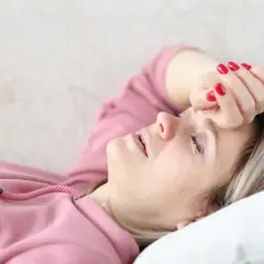 Kobieta w różowej bluzie leży na kanapie z ręką na czole, z zamkniętymi oczami.