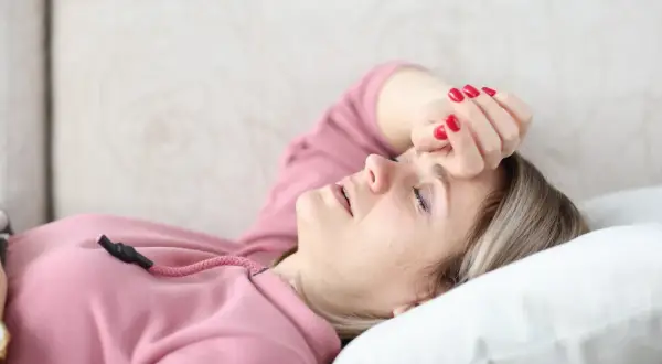 Kobieta w różowej bluzie leży na kanapie z ręką na czole, z zamkniętymi oczami.