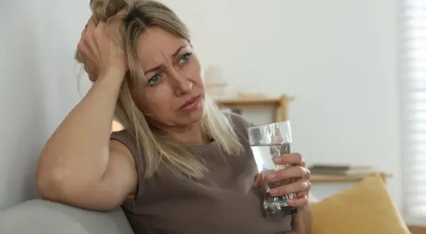 Kobieta w średnim wieku z ręką na głowie, trzymająca szklankę wody, z wyrazem zmartwienia na twarzy.
